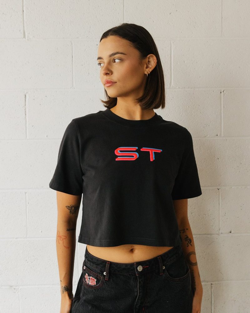 Model wearing the black Simply Techno ladies crop tee with the red and cyan ST logo, against a white brick wall